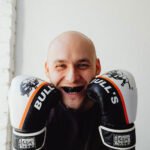 A man holding boxing gloves, smiling around a mouth guard