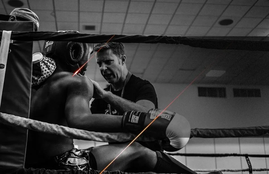 A black and white image of a coach talking to a young boxer, sat in the corner of a boxing ring